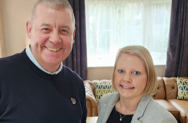 A man in a blue jumper smiling, with a women with blond hair, blue eyes, wearing a gray jacket smiling