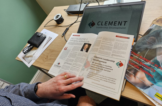 A hand holding open the pages of a magazine which is about coaching.  The magazine is resting on a desk near a computer.