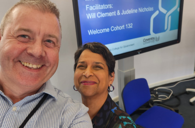 Smiling man in shirt Will and a woman with short dark heir with hoop earrings stood together with a projector screen in the back with text on