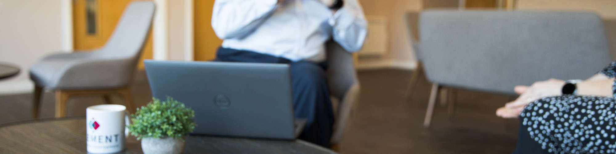 Man in a blue shirt talking to a woman.  There is a mug and notepad on a coffee table in front of them.