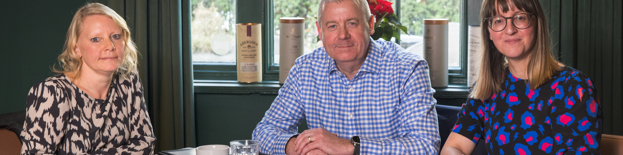 Three people sit at a wooden table in a café-style meeting space, looking at the camera. They have notebooks, coffee cups, and glasses of water in front of them. Behind them is a large window with green curtains and several boxed bottles on the sill, with
