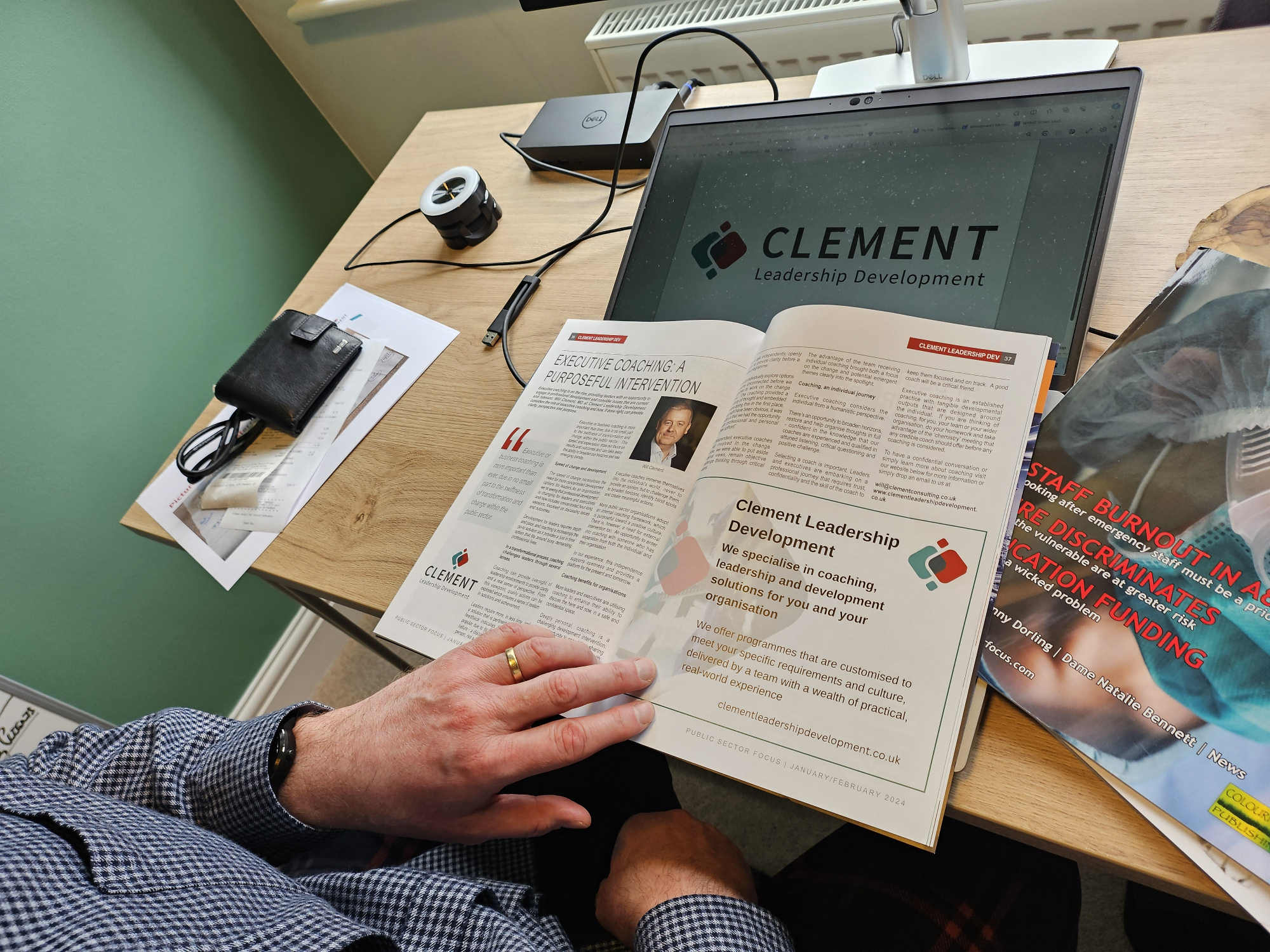 A hand holding open the pages of a magazine which is about coaching.  The magazine is resting on a desk near a computer.