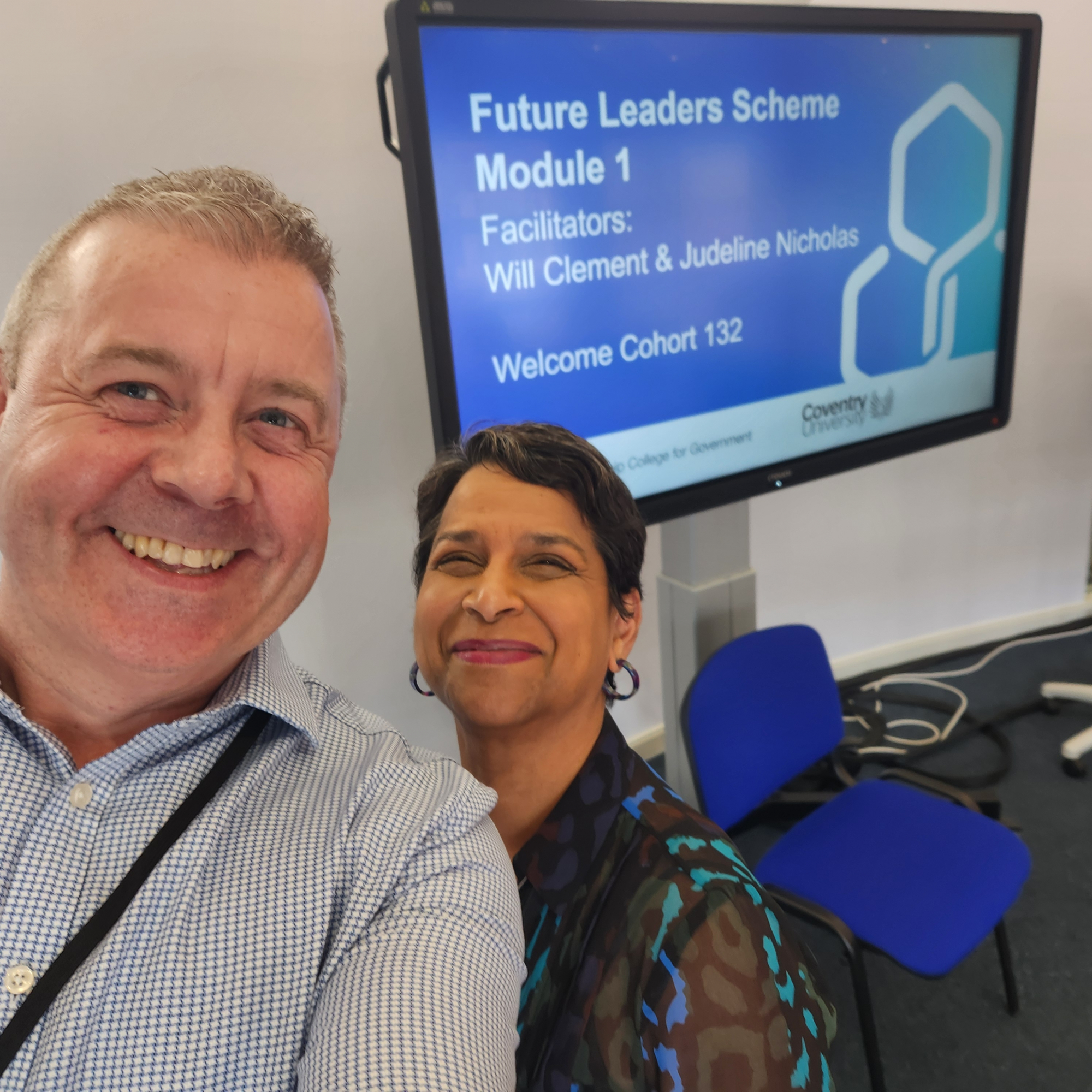 Smiling man in shirt Will and a woman with short dark heir with hoop earrings stood together with a projector screen in the back with text on